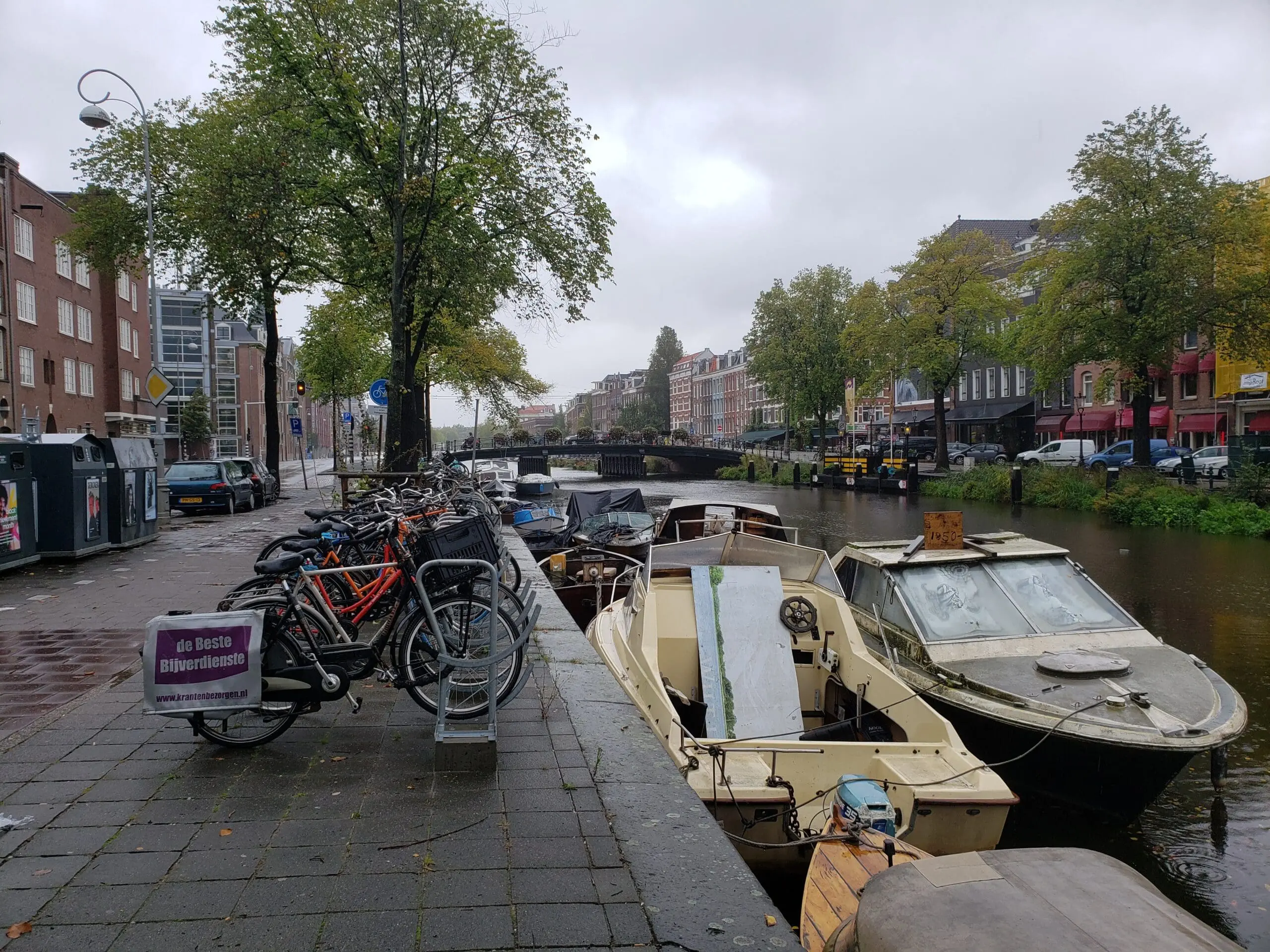 Motorboats tied along an Amsterdam canal beside a long bike rack, with trees, parked boats, and a distant bridge with flowers.