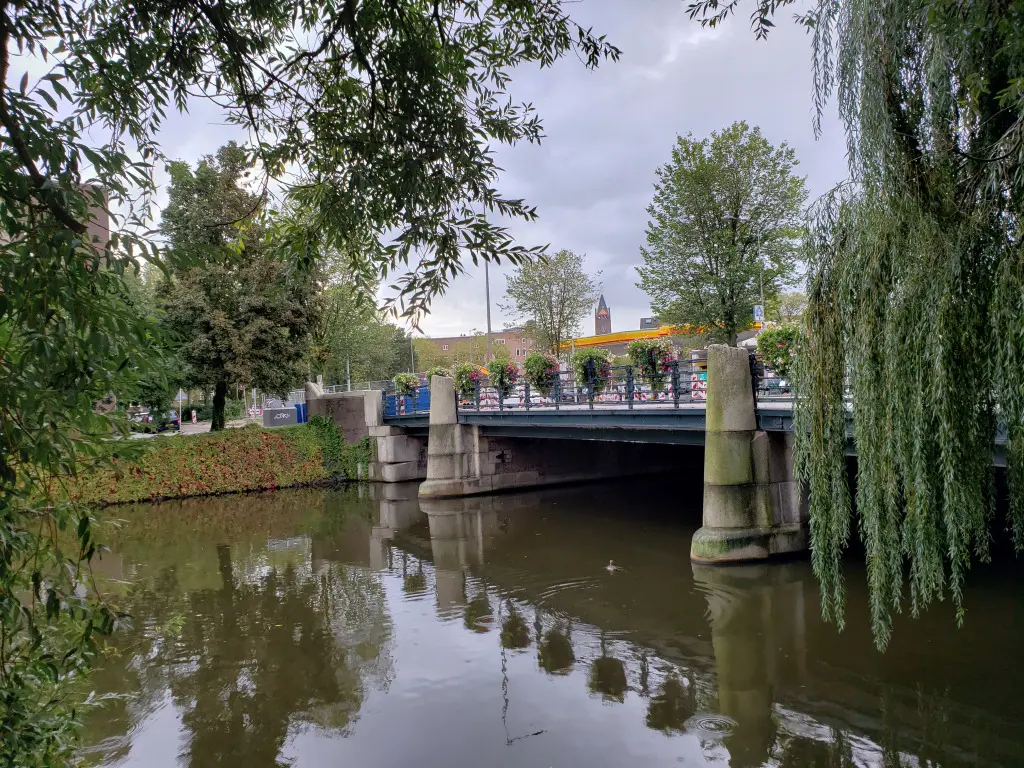 Dark green Amsterdam canal with willow branches, a flower‑lined bridge, dense trees, and a duck swimming toward the shadows.