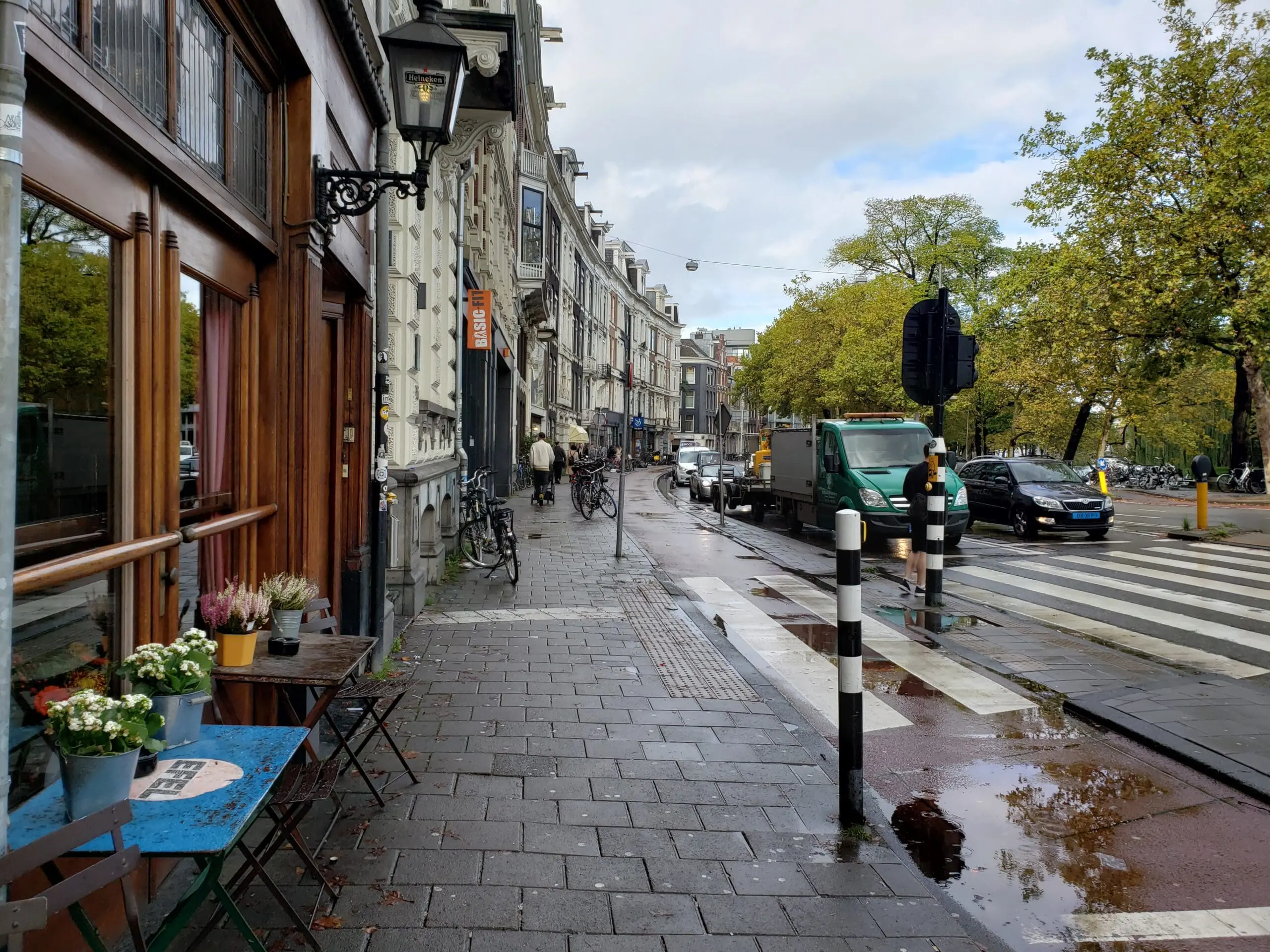 Amsterdam street with a wet brick sidewalk, older buildings, café tables, bicycles, and golden autumn trees under an overcast sky.