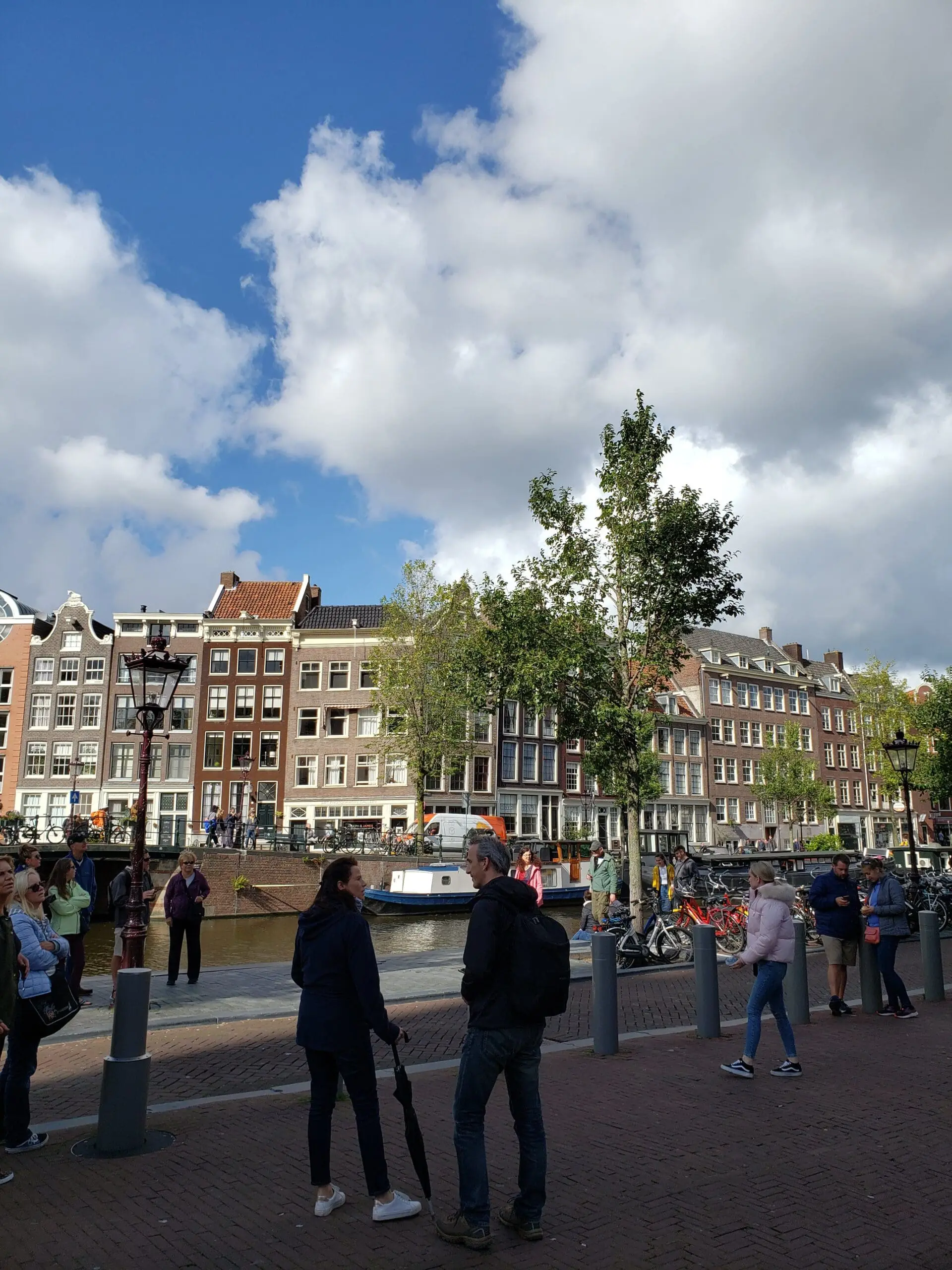 People walking along a worn red‑brick Amsterdam sidewalk beside a canal, with old buildings, bicycles, streetlamps, and young green trees.
