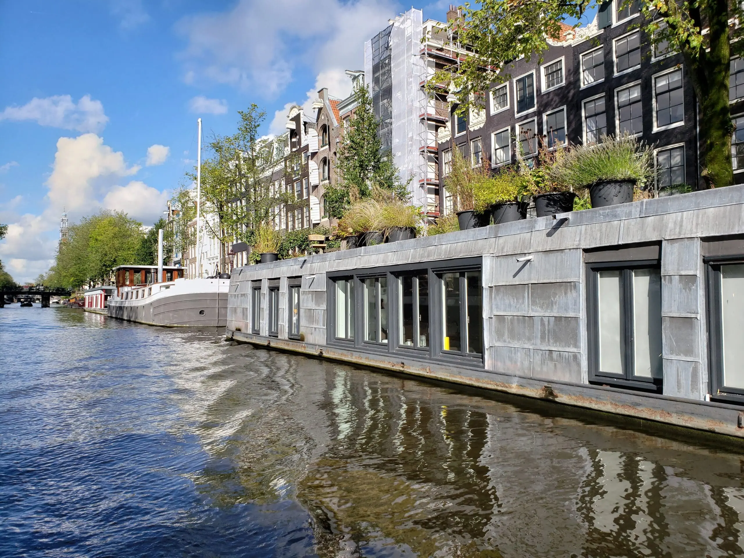 Long cement houseboat on an Amsterdam canal with sharp angles leading to a distant vanishing point, reflected in rippling water beneath blue sky and white clouds.
