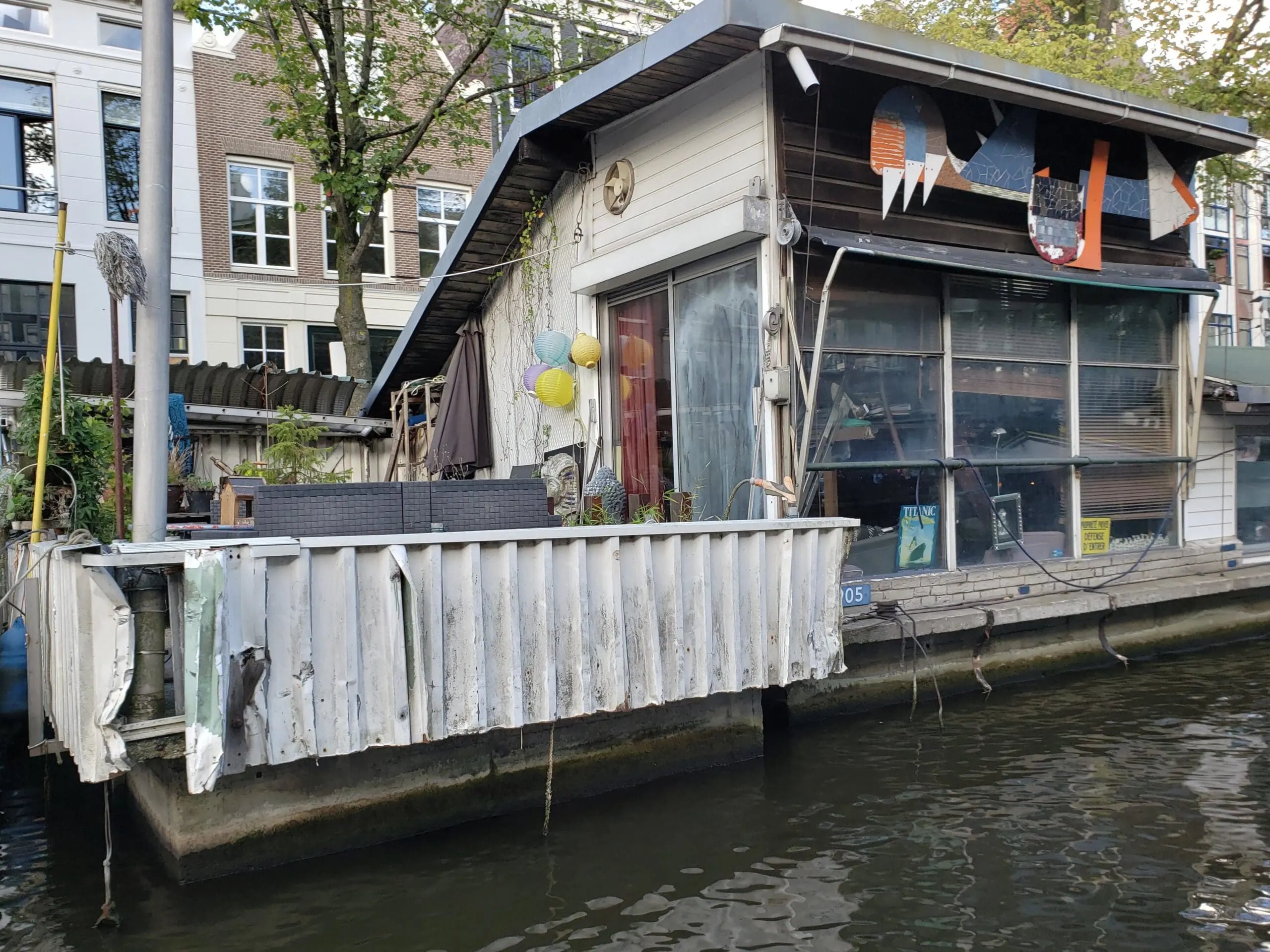 Old weathered houseboat on a dark Amsterdam canal with grey siding, worn windows, and a floating‑bar look.