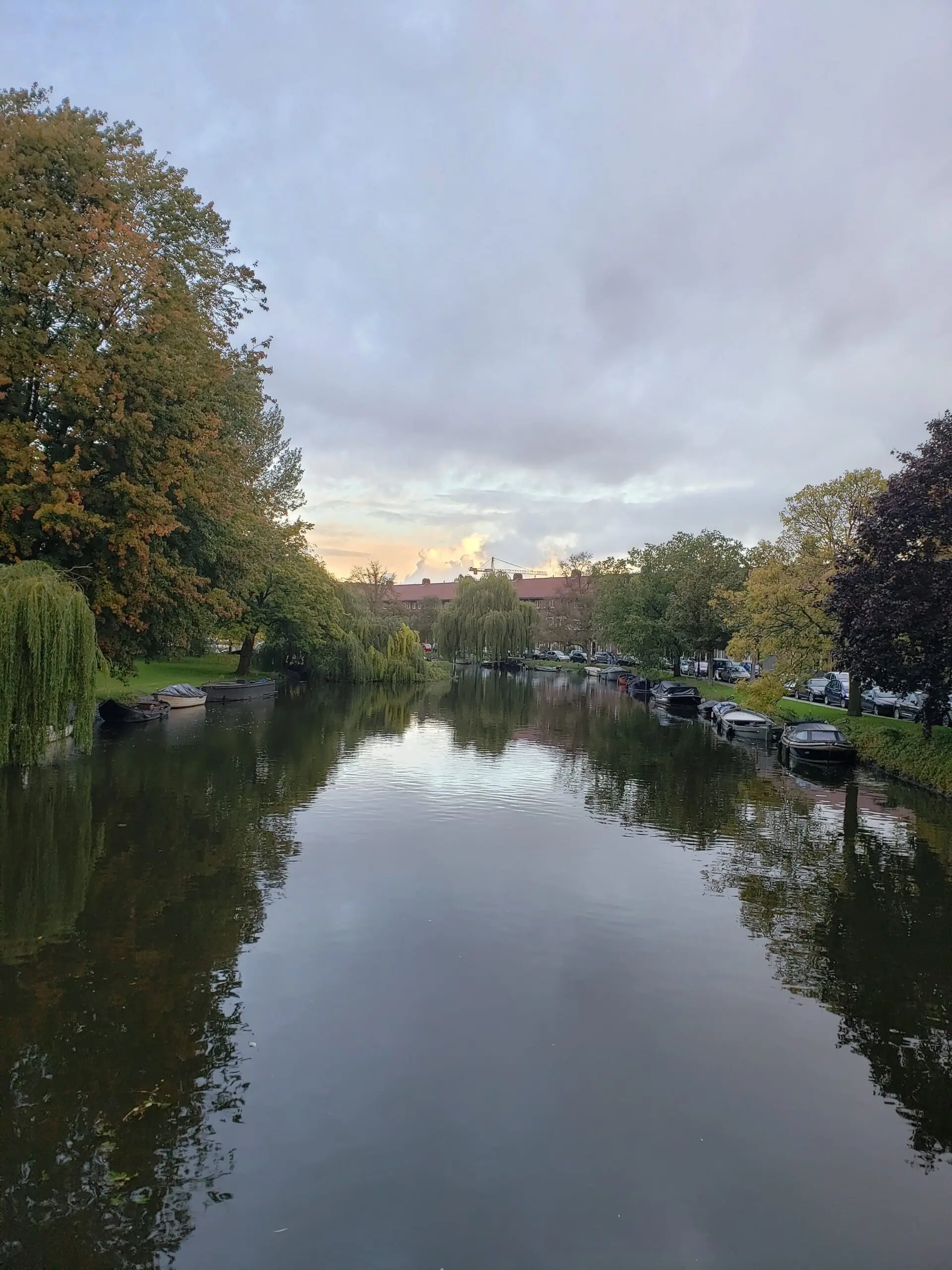 View from a small Amsterdam bridge overlooking a canal lined with motorboats, autumn trees, grasses, and a bright reflection under an overcast sky.