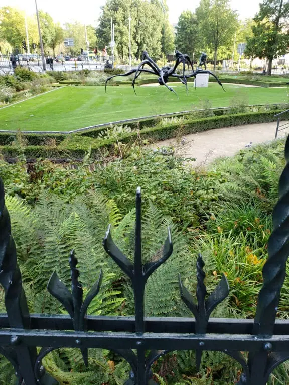 Wrought‑iron spider sculpture behind a trident‑topped fence, surrounded by water, grass, and city trees on a sunny afternoon in Amsterdam.