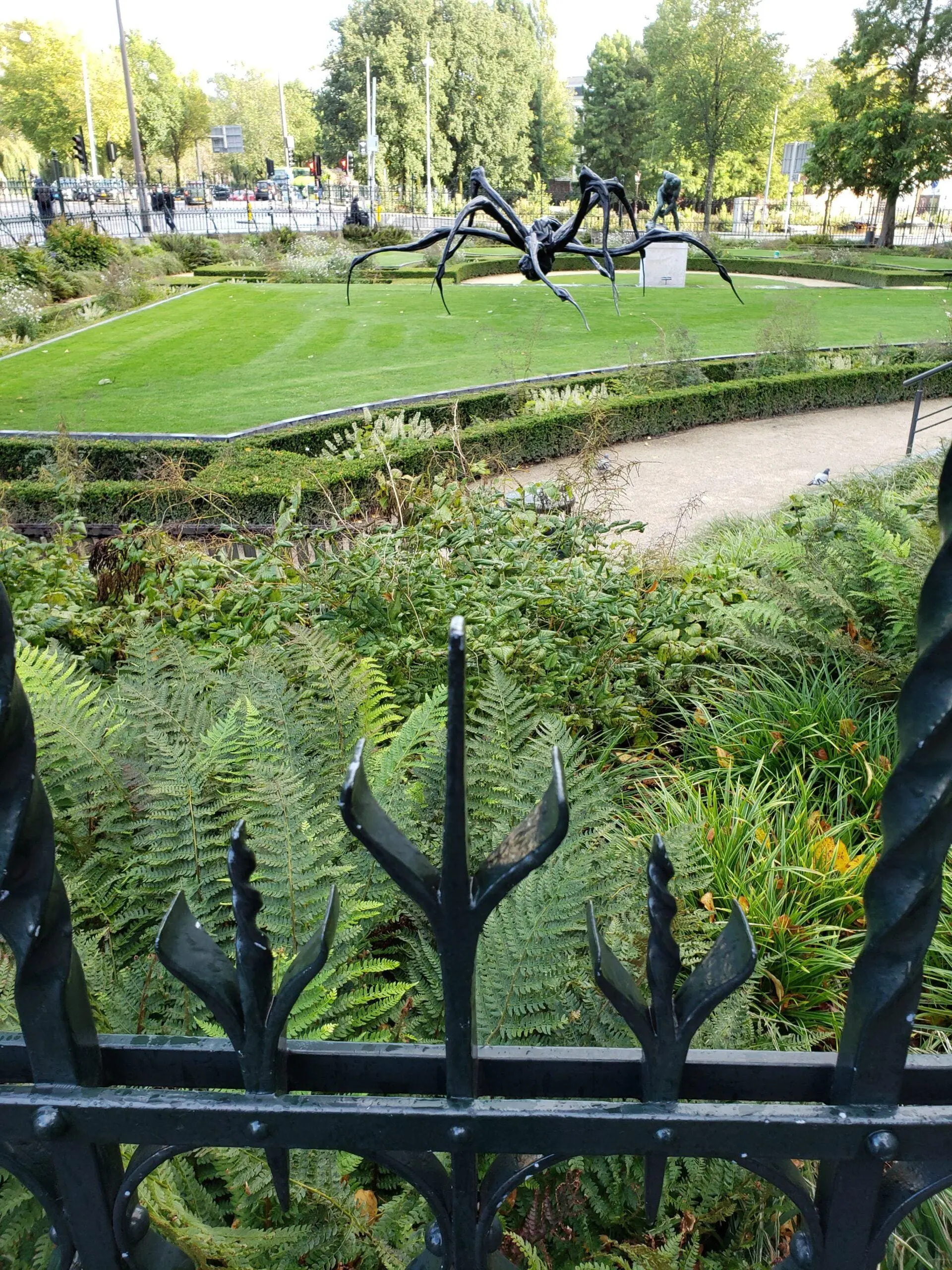 Wrought‑iron spider sculpture behind a trident‑topped fence, surrounded by water, grass, and city trees on a sunny afternoon in Amsterdam.
