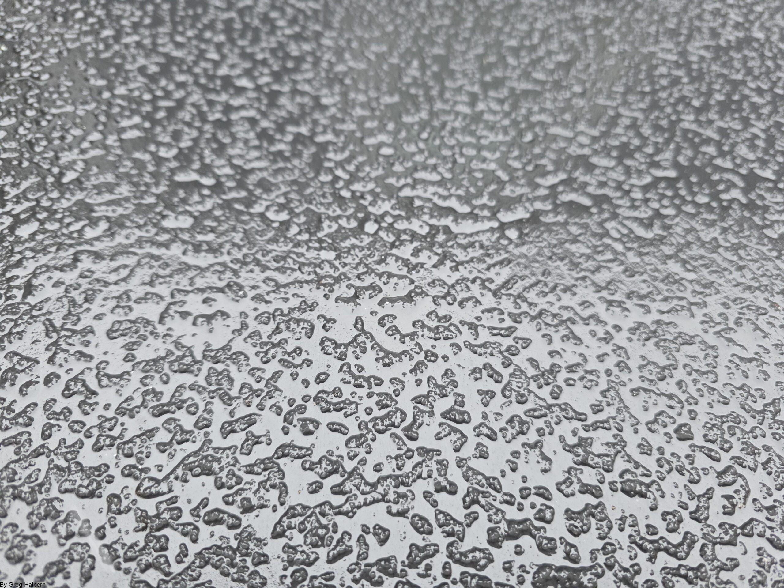 Rain droplets forming textured patterns on a black metal surface with tonal bands and squared-off illusion.