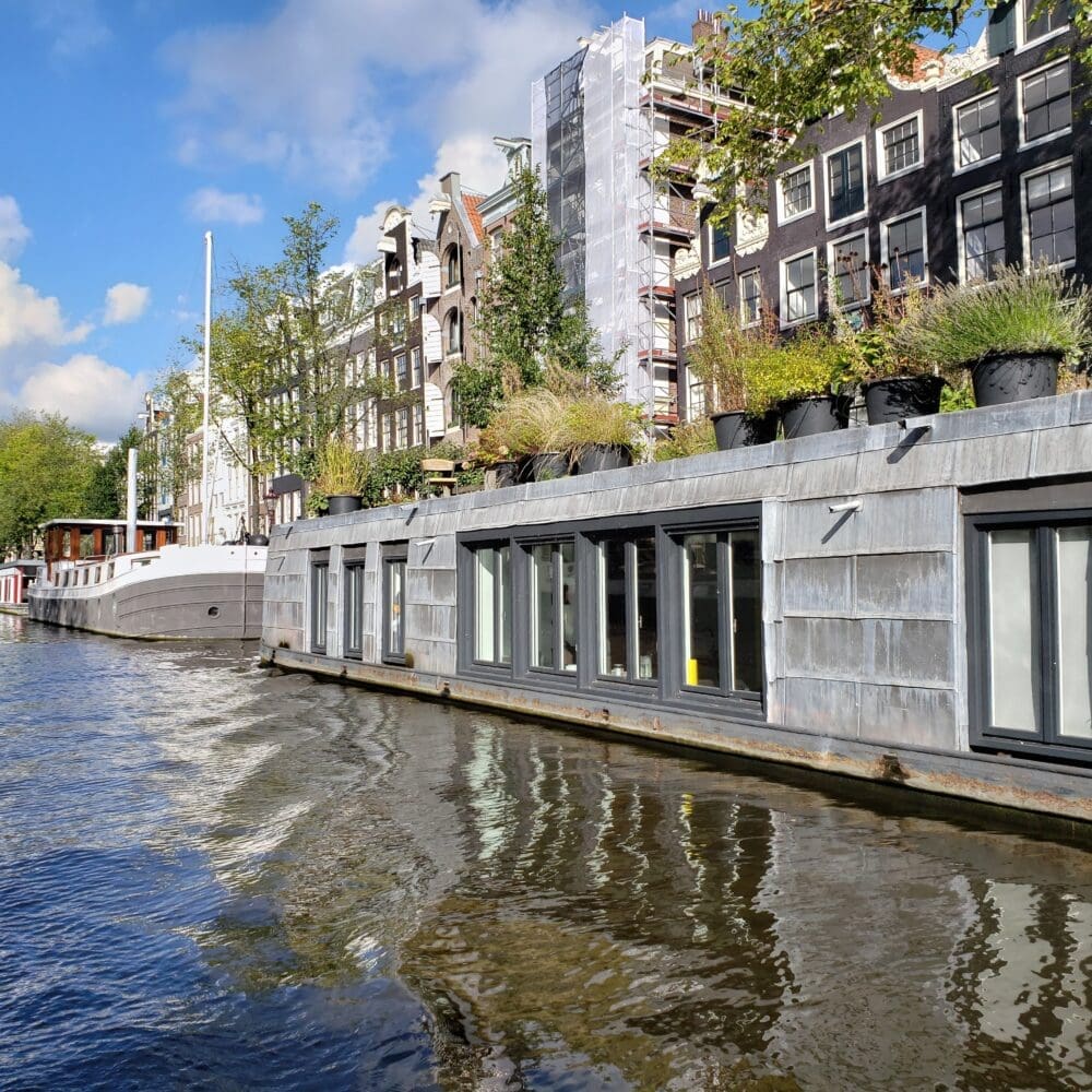 Long cement houseboat on an Amsterdam canal with sharp angles leading to a distant vanishing point, reflected in rippling water beneath blue sky and white clouds.
