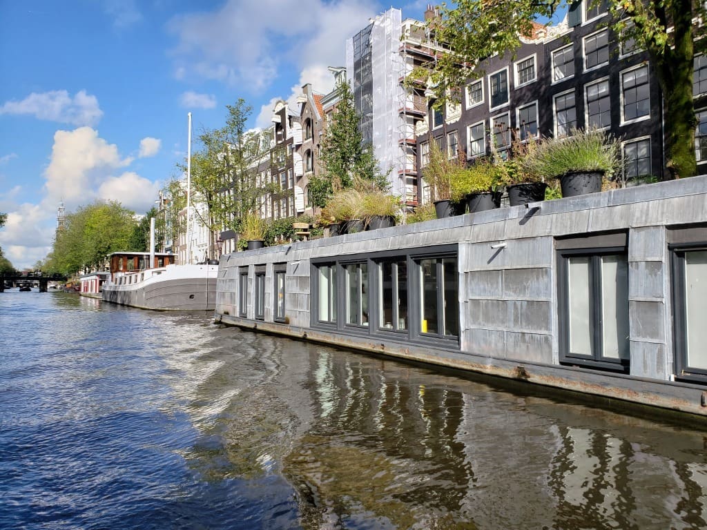 Sunny day on an Amsterdam canal. puffy clouds splash the perfect blue sky. on the side of the canal is a very long house boat, one of a permanent set of water-bound residences. there's a bridge in the distance and other houseboats lining the bank.