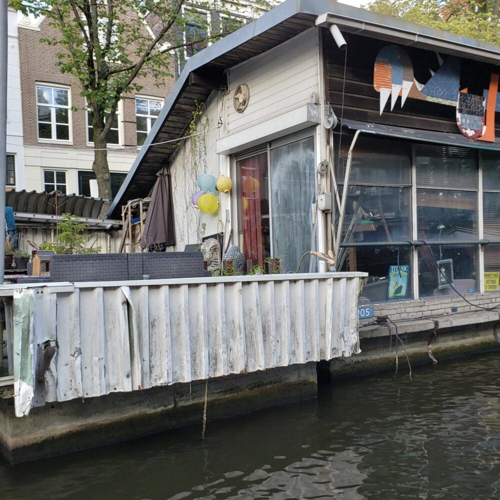 Ramshackle Amsterdam houseboat with corrugated siding, mismatched wood panels, and large glass windows floating on a dark canal.