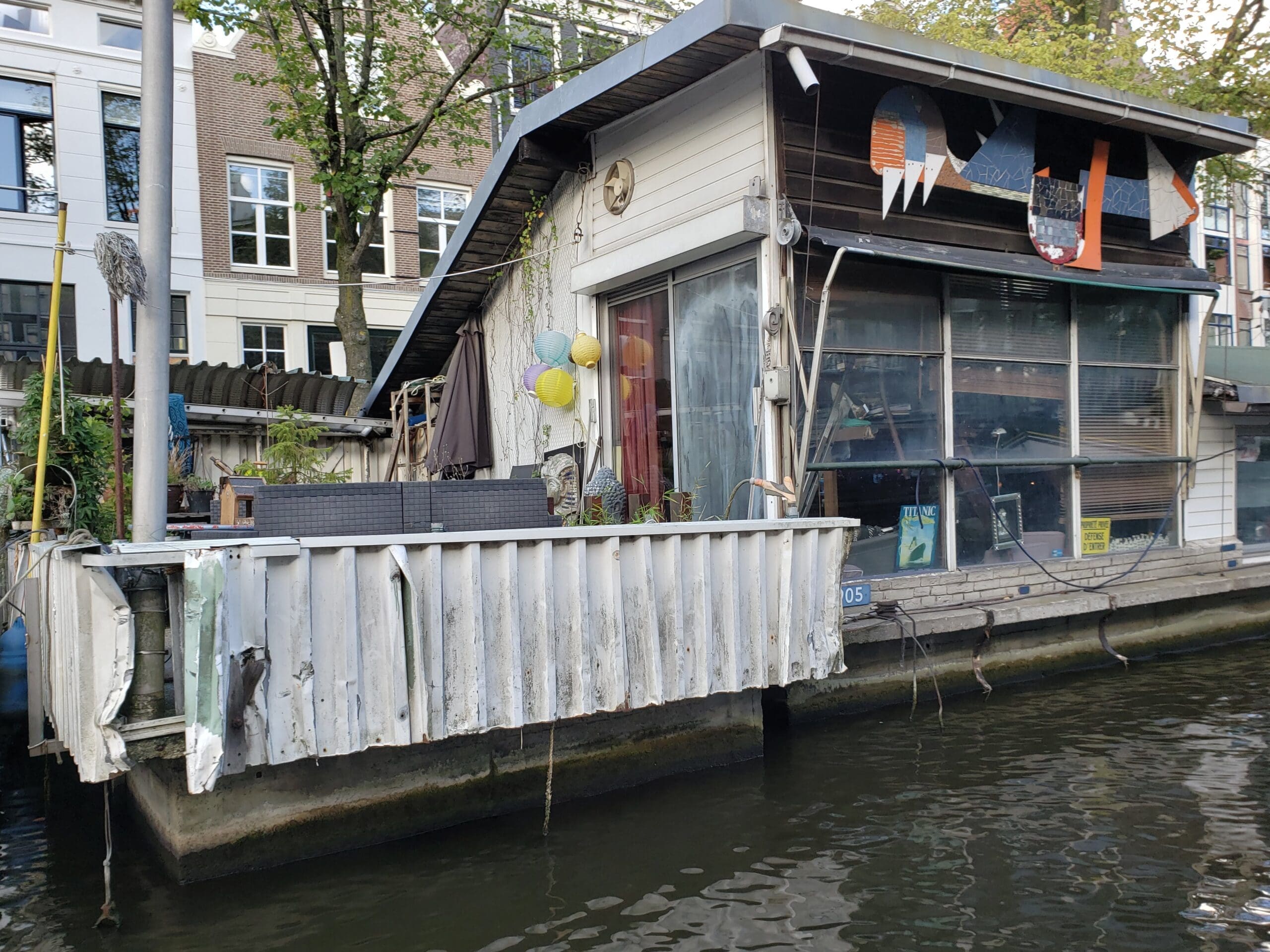 Ramshackle Amsterdam houseboat with corrugated siding, mismatched wood panels, and large glass windows floating on a dark canal.