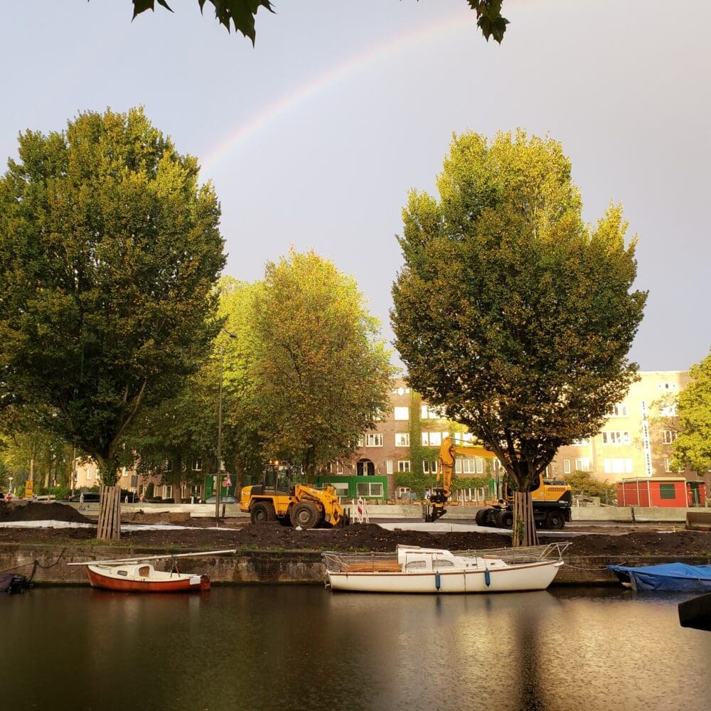 Rainbow over an Amsterdam canal after rain, with trees, motorboats, and orange construction vehicles along the shore.