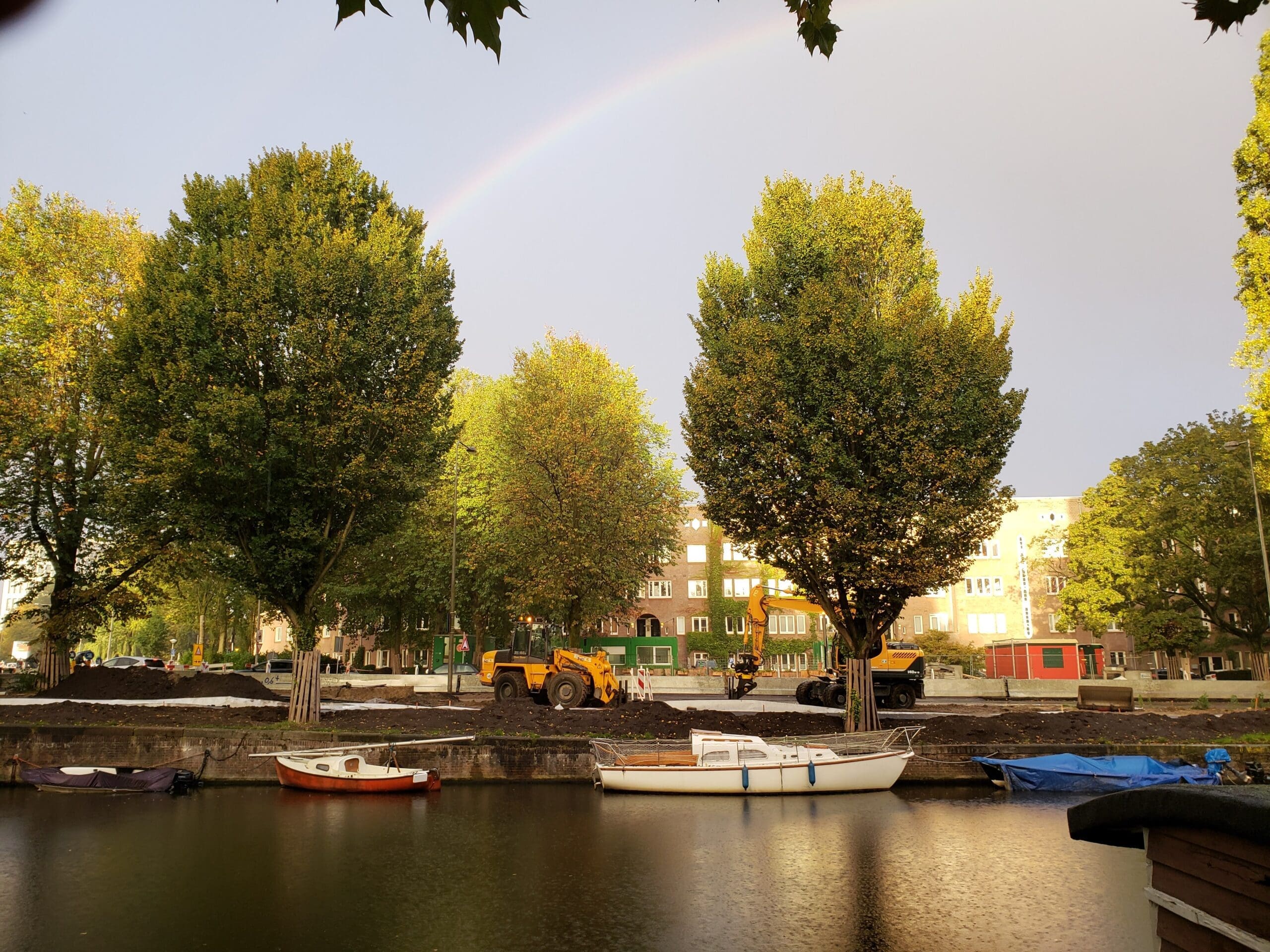 Rainbow over an Amsterdam canal after rain, with trees, motorboats, and orange construction vehicles along the shore.