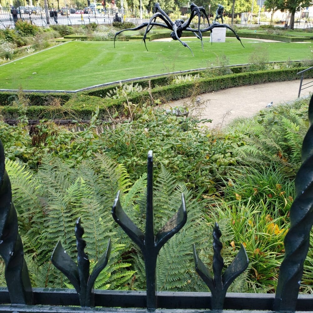 Wrought‑iron spider sculpture behind a trident‑topped fence, surrounded by water, grass, and city trees on a sunny afternoon in Amsterdam.